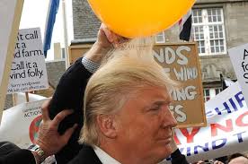 A close-up photo of Donald Trump with a yellow balloon held above his head, partially obscuring his hair. He is surrounded…