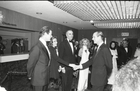 A black-and-white photo shows two men in tuxedos shaking hands in a formal setting, possibly a theater lobby, with a…
