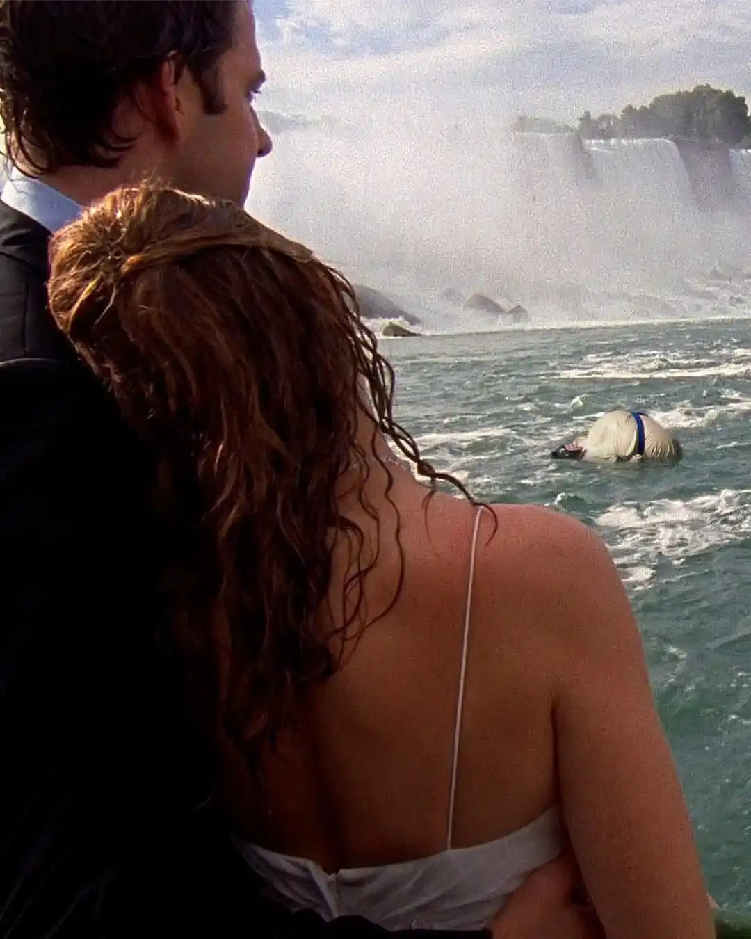 A man and woman in formal attire stand close together, looking out at a large waterfall. In the water below them, a person…