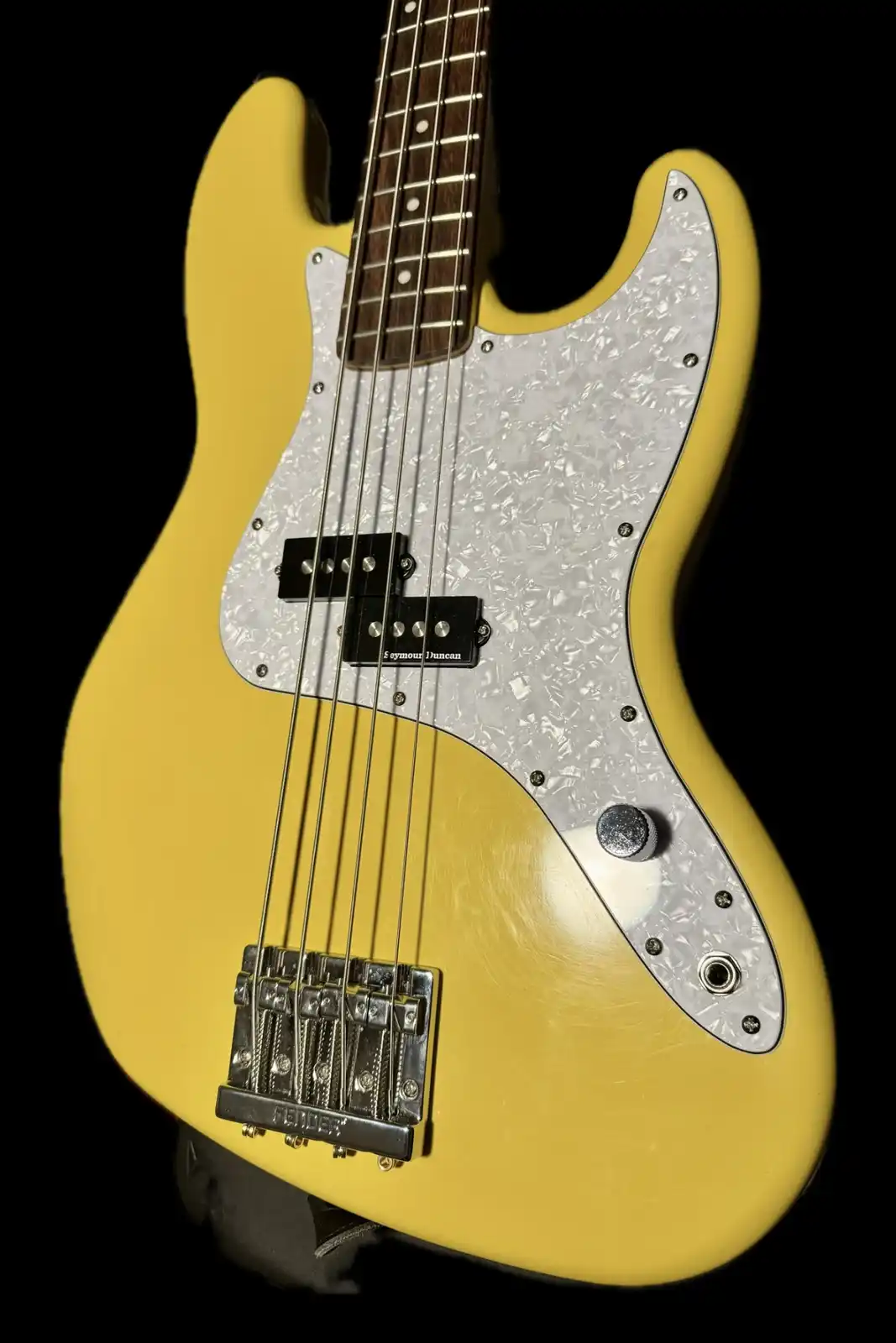 A close-up photo of a yellow Fender bass guitar with a white pickguard and four strings, set against a black background. The…