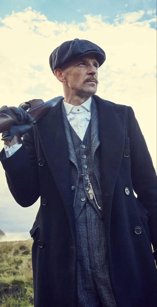 A man in a dark coat, vest, and flat cap stands outdoors holding a shotgun over his shoulder, looking upward with a serious…