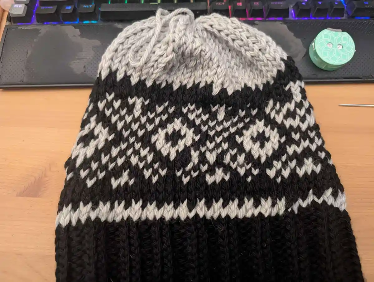 A hand-knitted beanie with a black and gray patterned design sits on a wooden desk in front of a backlit keyboard. The hat…