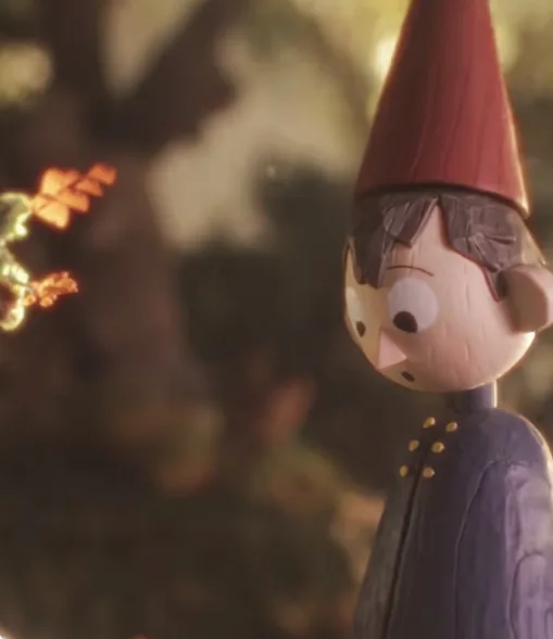 A close-up of a wooden doll with a red cone hat, wide eyes, and a surprised expression, standing in a blurry forest setting…