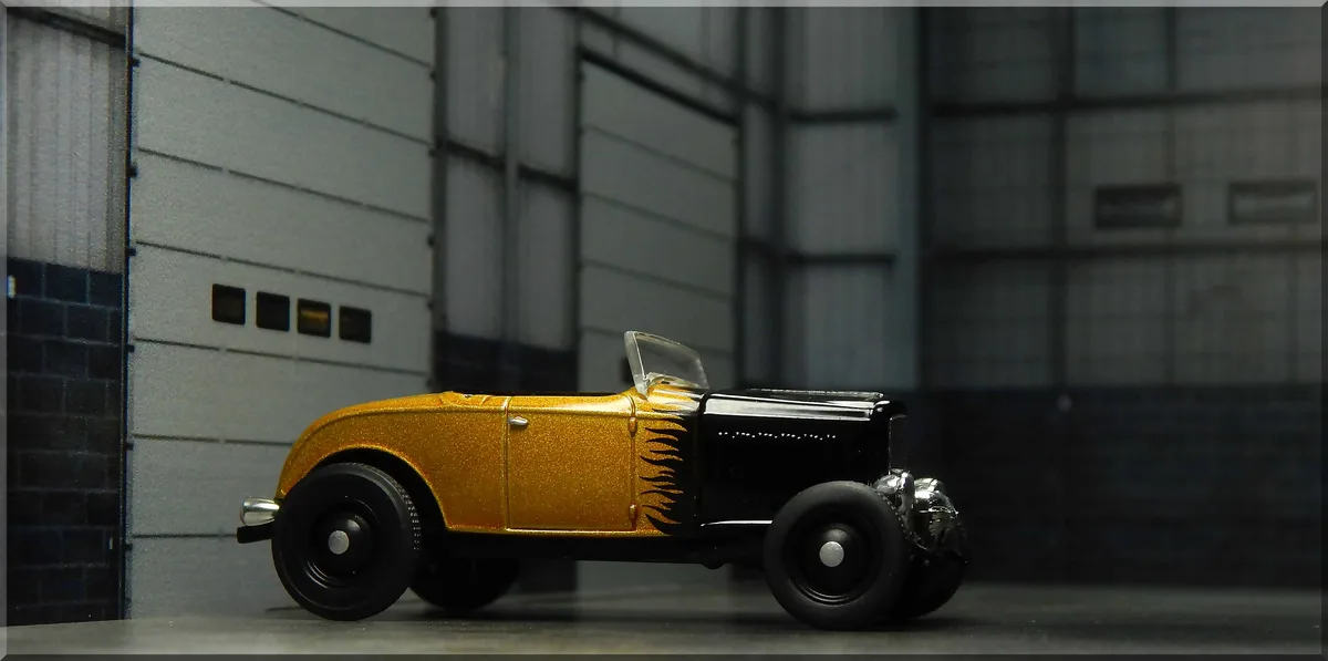 A gold and black toy hot rod model with flame decals is shown in profile against a gray industrial-style wall. The overlaid…