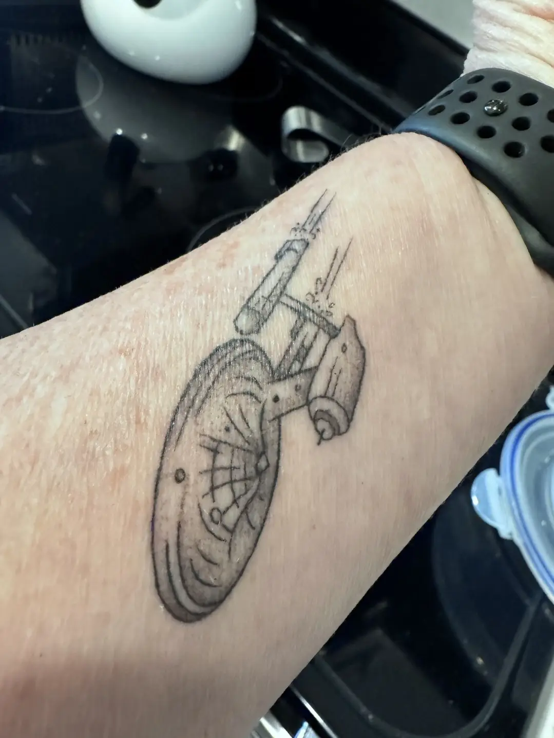 A close-up photo shows a black-and-white tattoo of a Starfleet ship, resembling the USS Enterprise, on a person's forearm…