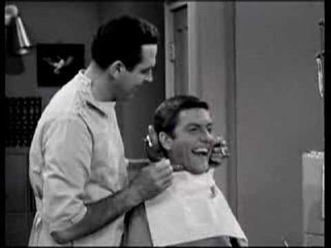a black-and-white still from an old TV show shows a barber holding clippers to a customer's neck while the customer laughs…