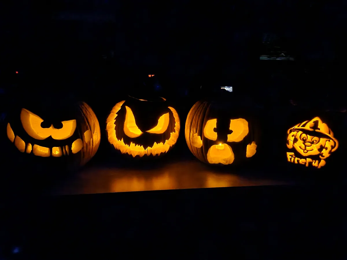 Four carved pumpkins glow in the dark, each with a different face: three menacing jack-o'-lanterns and one depicting a…
