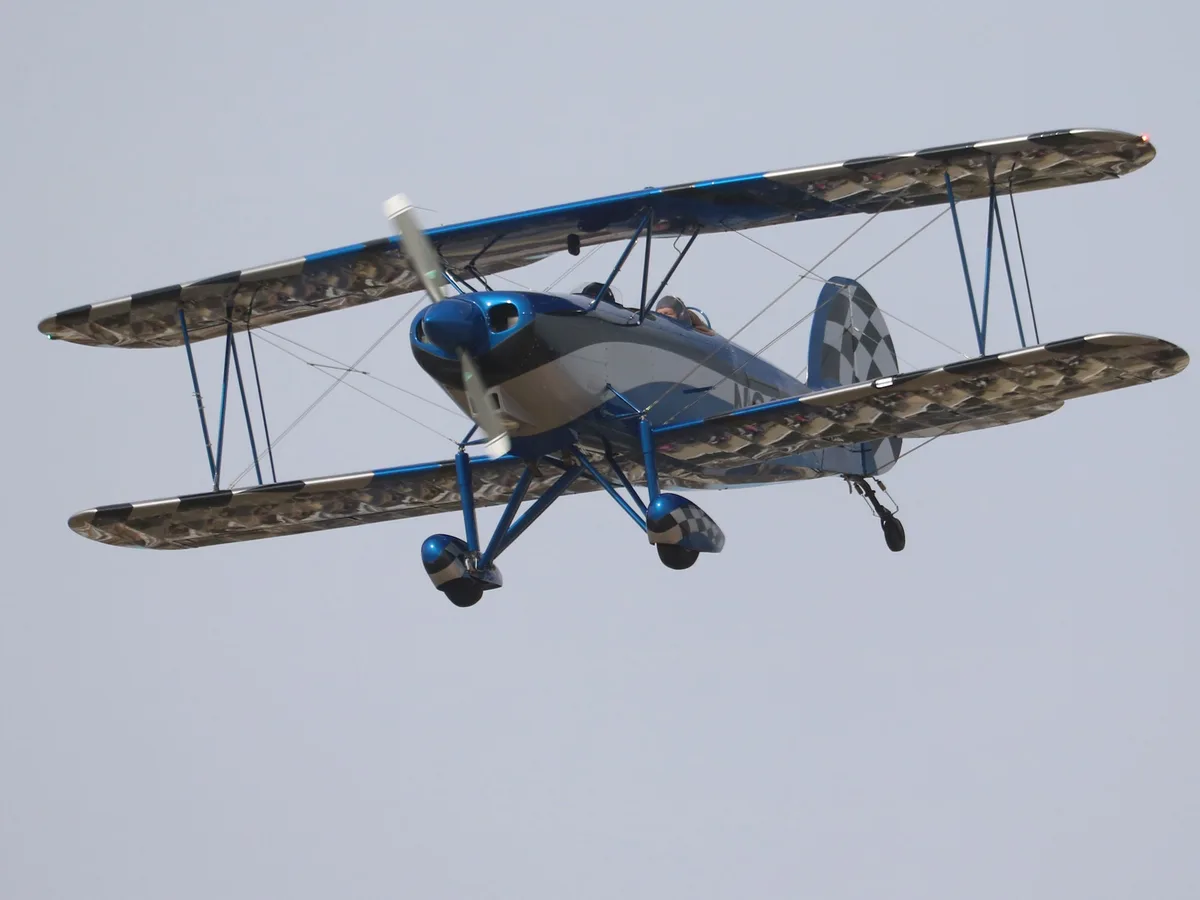 A blue and silver biplane with checkered patterns on its wings, tail, and wheels is flying against a pale sky. The pilot is…