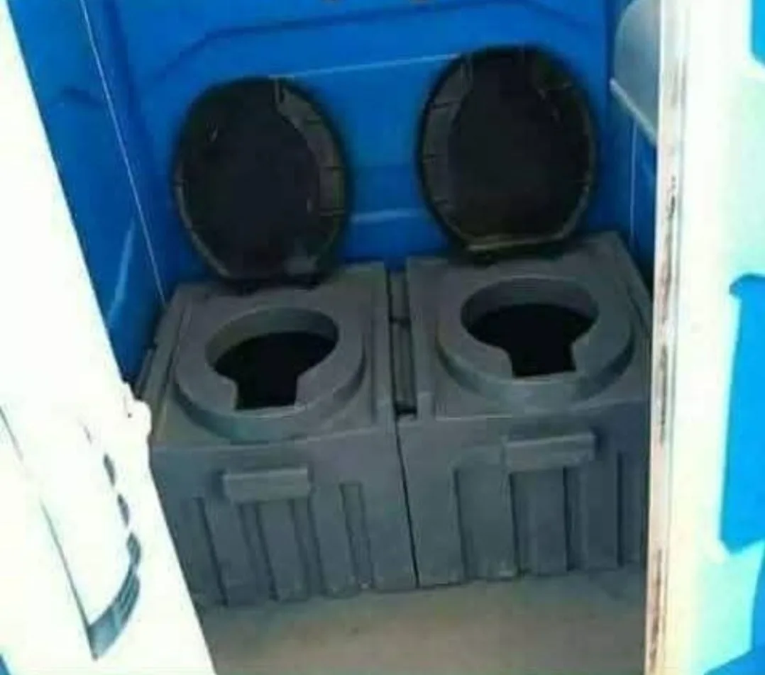 The image shows two side-by-side portable toilets inside a blue enclosure, both with their black plastic lids lifted open…