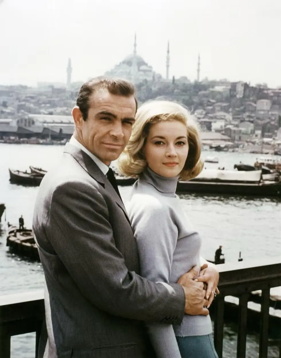 a vintage promotional photo shows actor sean connery in a grey suit embracing a blonde woman in a light grey turtleneck…