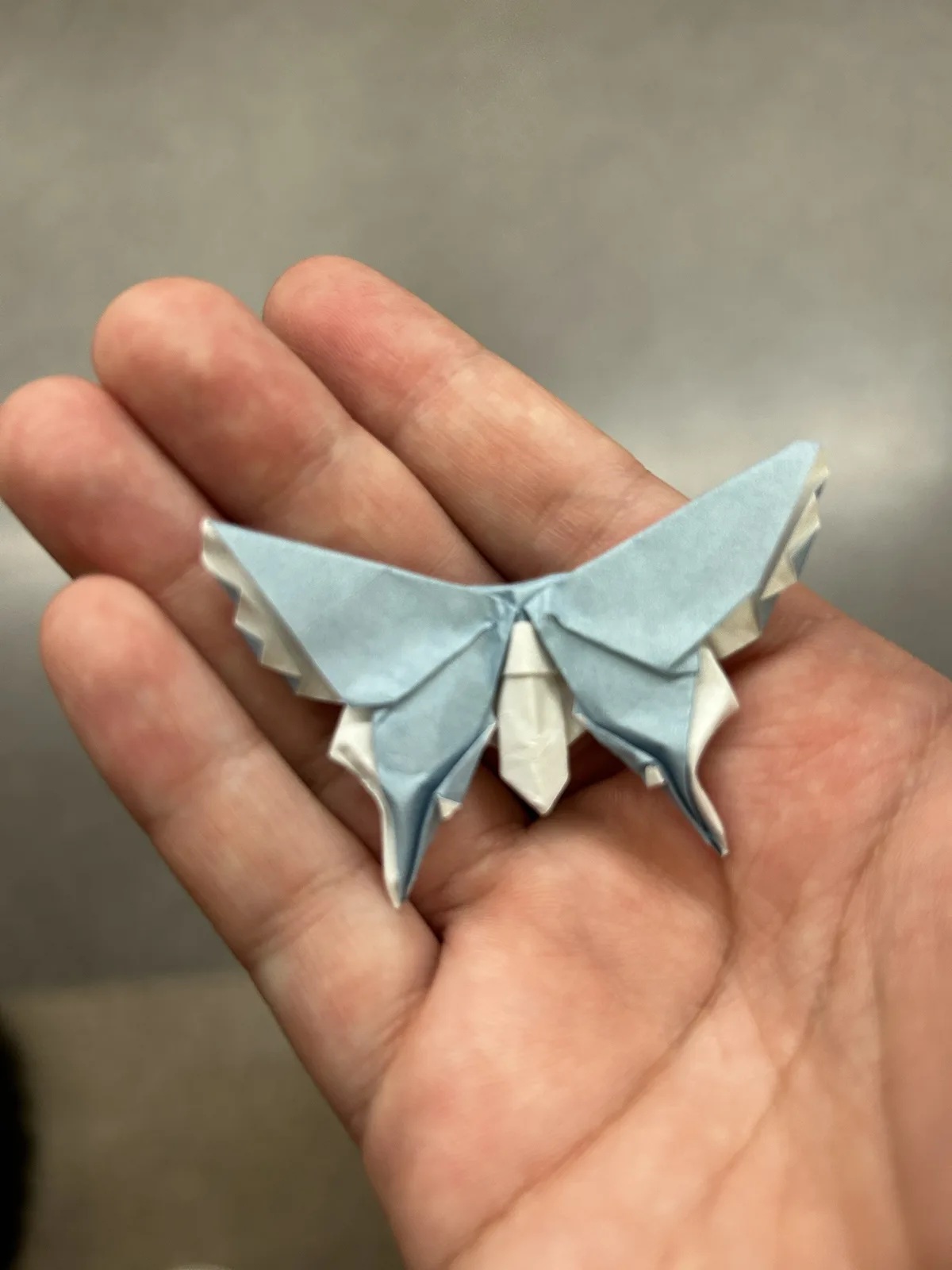 A hand holds a delicate origami butterfly made from light blue and white paper, showcasing intricate folds and realistic…