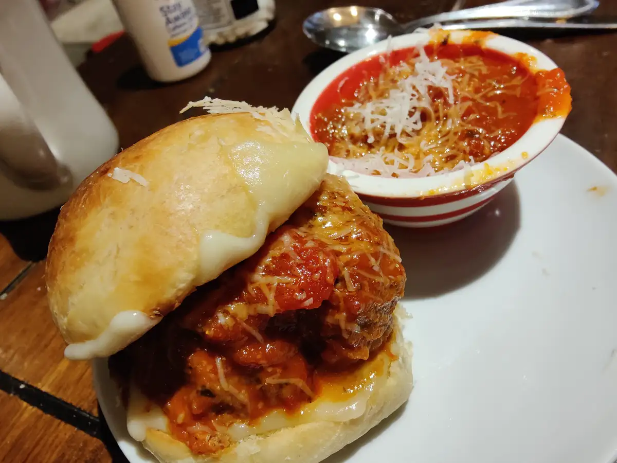 A close-up photo shows a meatball sub on a white plate, with melted cheese oozing from the bun and a side bowl of tomato…