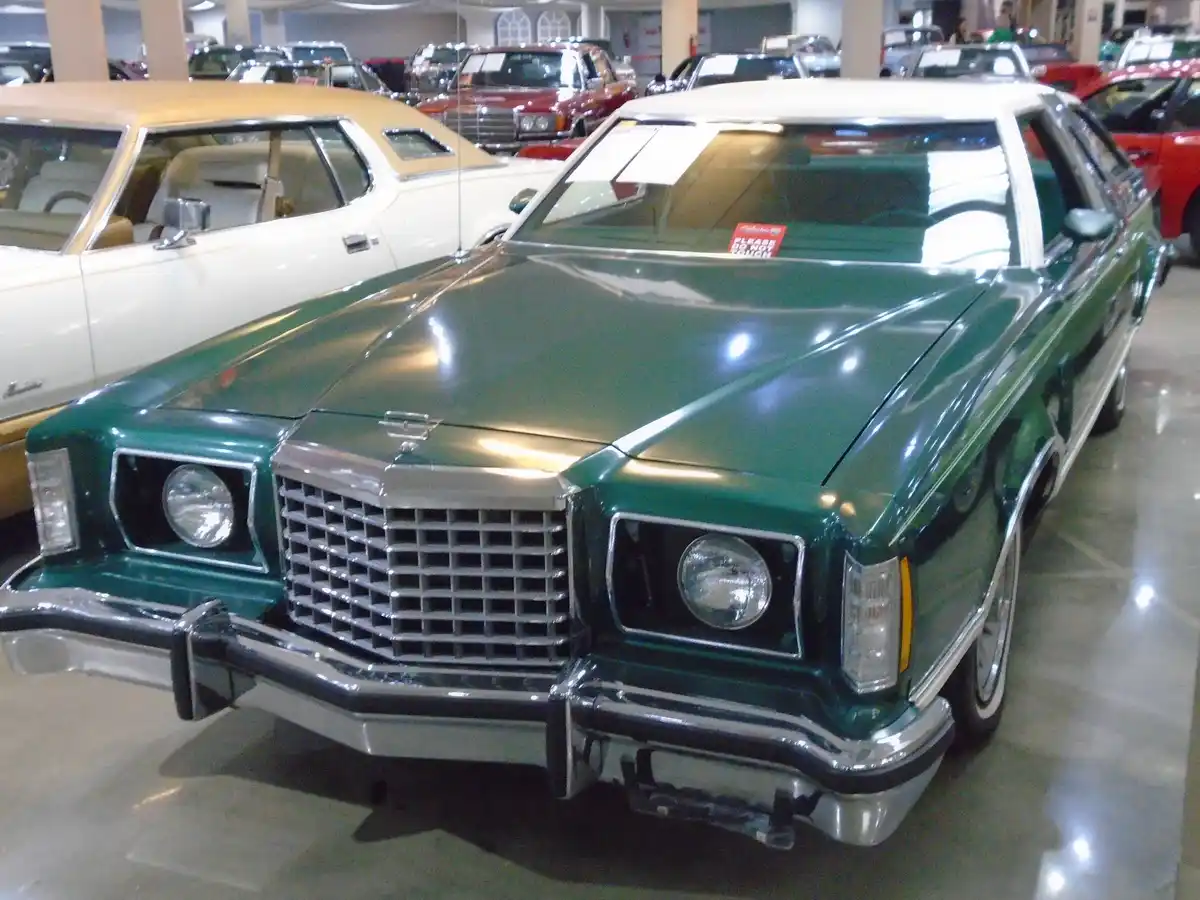 A shiny green vintage Cadillac sedan is displayed in a car museum, with a small red sign on its windshield that reads…