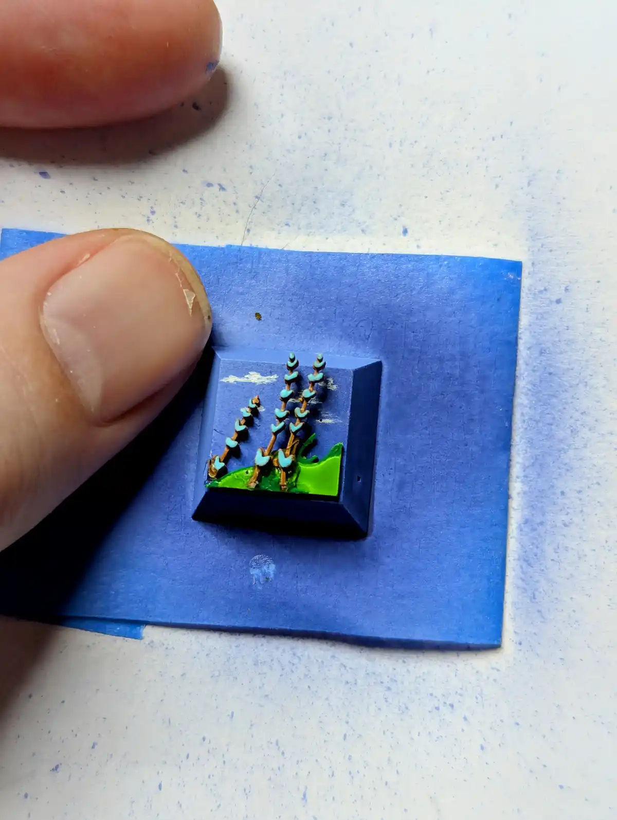 A close-up photo shows two fingers holding a small, square, dark blue keyboard key. The key has been painted to resemble a…