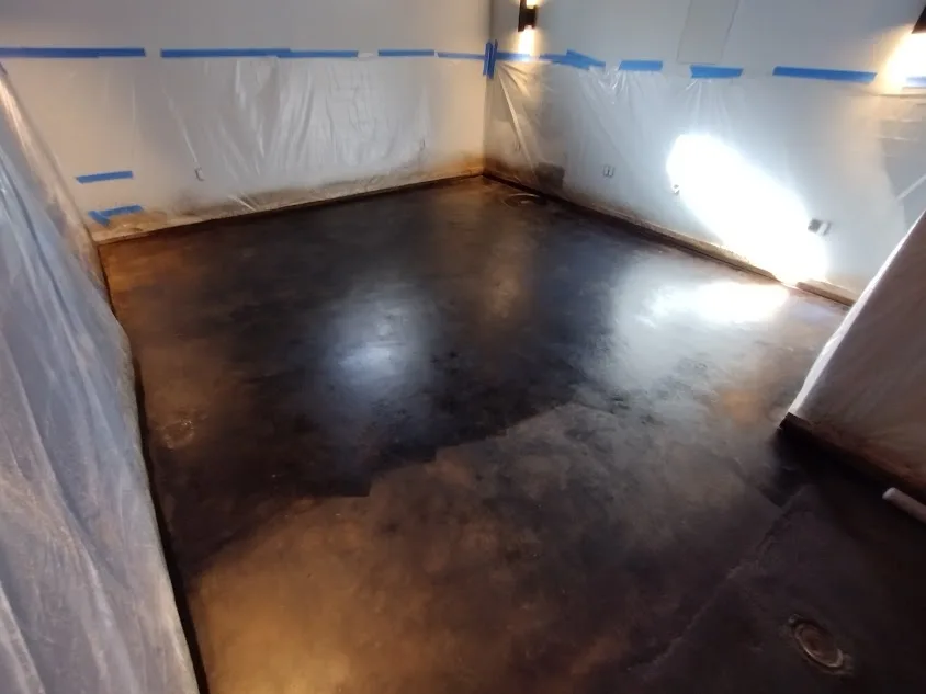 A room with a dark, stained concrete floor and walls covered in white plastic sheeting secured with blue painter's tape…