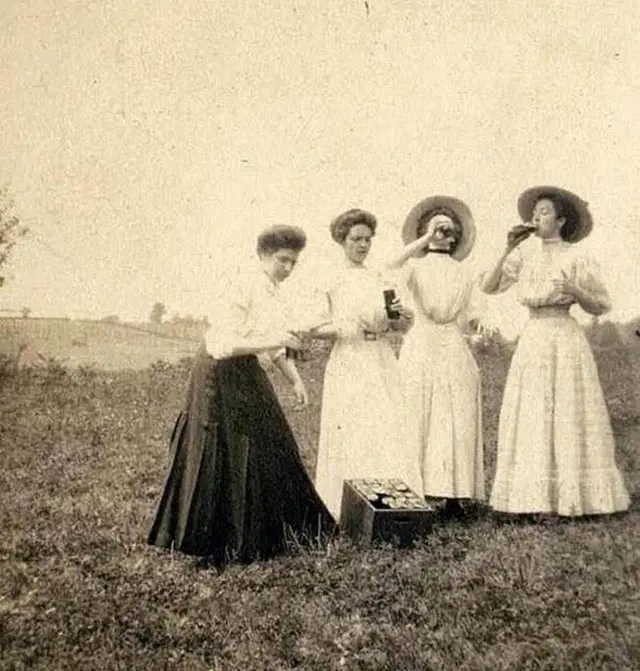 A sepia-toned vintage photograph shows four women in long dresses and hats outdoors on grass. One woman on the left is…