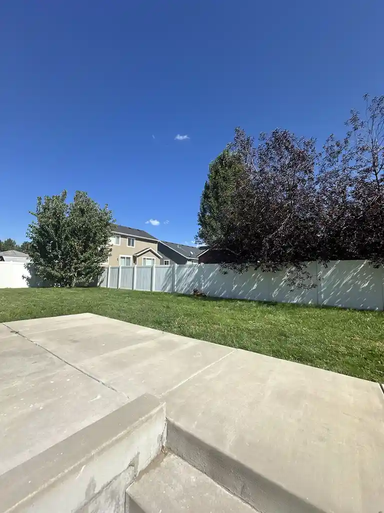 A low-angle photo shows a concrete patio with steps leading up to a grassy backyard bordered by a white fence, under a clear…