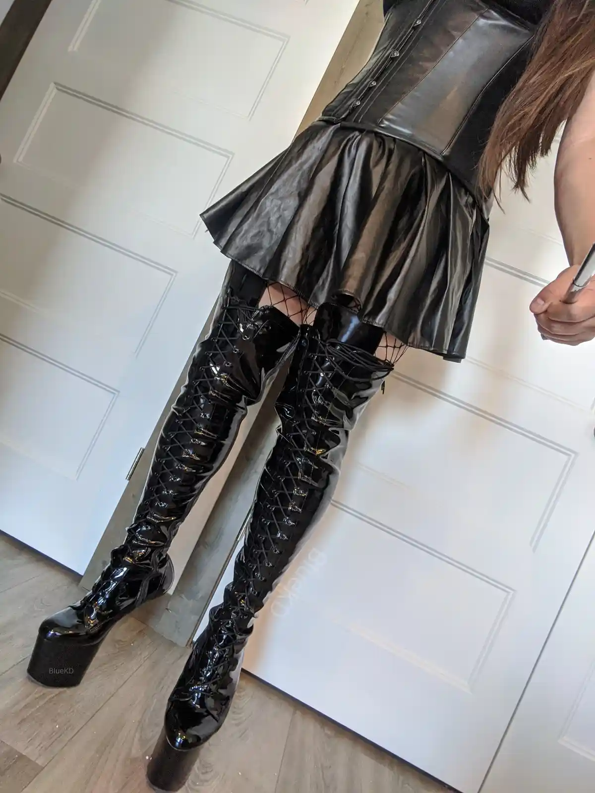A person wearing a shiny black corset and matching pleated skirt stands indoors, their legs covered in tall, glossy black…
