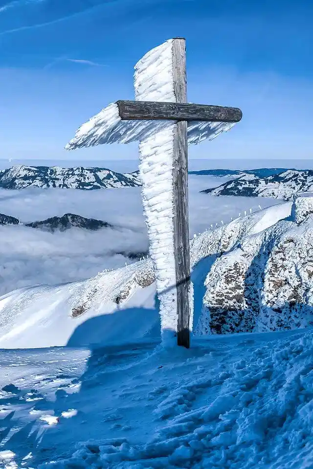 A wooden cross stands atop a snowy mountain peak, heavily coated in ice and snow, with a sea of clouds below and distant…