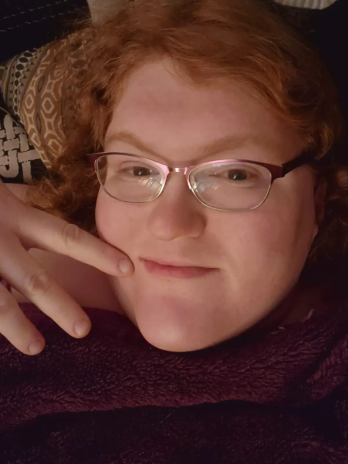 A person with curly red hair and glasses is lying down, resting their chin on their hand, wearing a dark purple sweater. The…