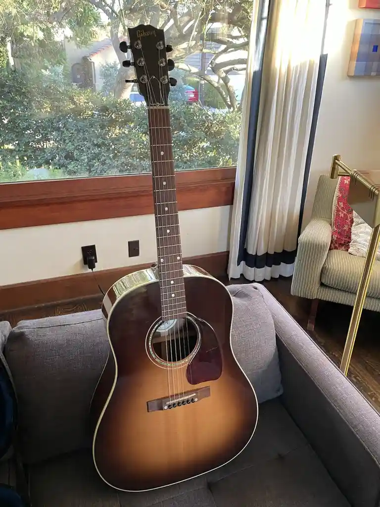 A Gibson acoustic guitar with a sunburst finish is propped upright on a gray couch in a sunlit living room, with a window…