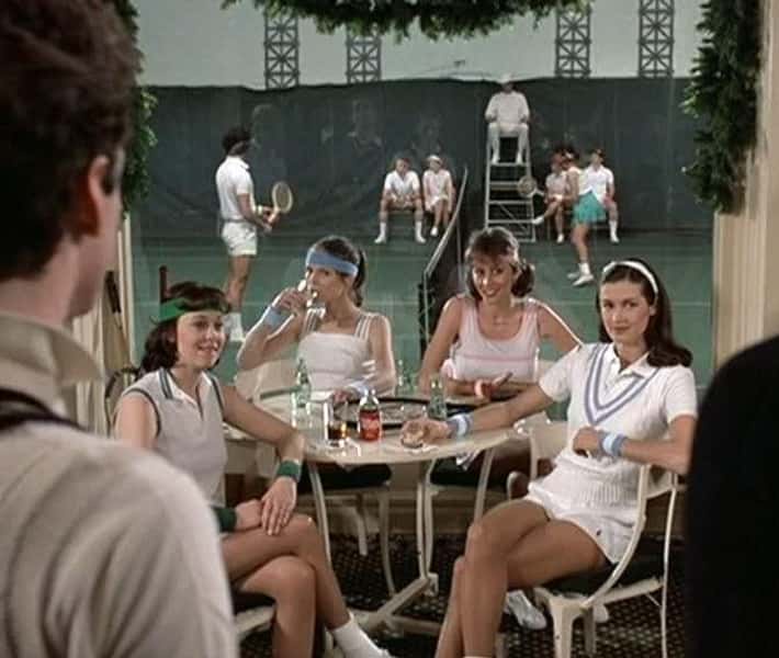 A still from a 1970s tennis movie shows four women in vintage tennis outfits sitting at a table, smiling and drinking. In…
