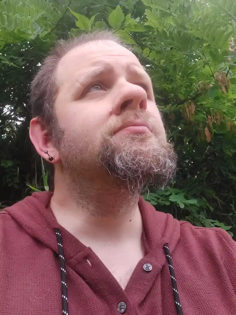 A man with a beard and ear piercing, wearing a maroon hoodie, looks upward with a slightly confused or contemplative…