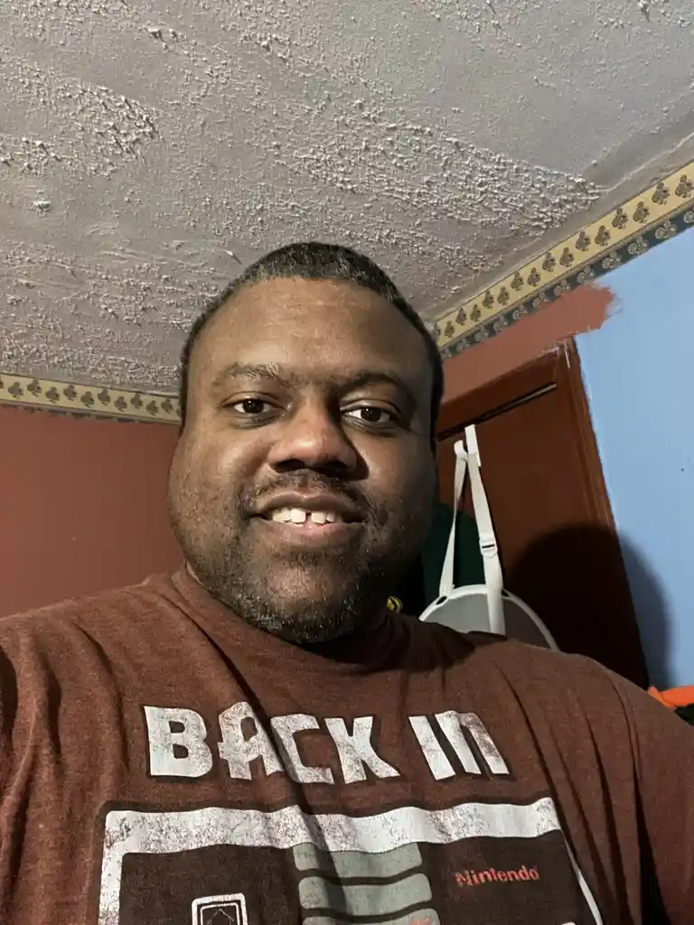 A man with a tooth missing smiles at the camera while wearing a brown t-shirt that reads 'BACK IN' with a Nintendo logo…