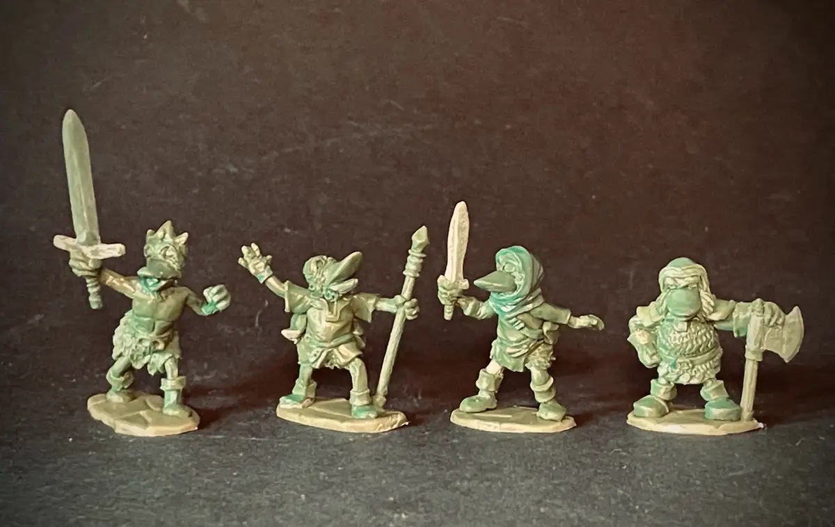 Four unpainted green miniature fantasy figures, resembling goblins or orcs, stand in a row on a dark surface. Each holds a…