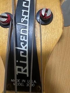 A close-up photo of a Rickenbacker bass guitar's headstock, showing the brand name in reverse lettering, with two tuning…