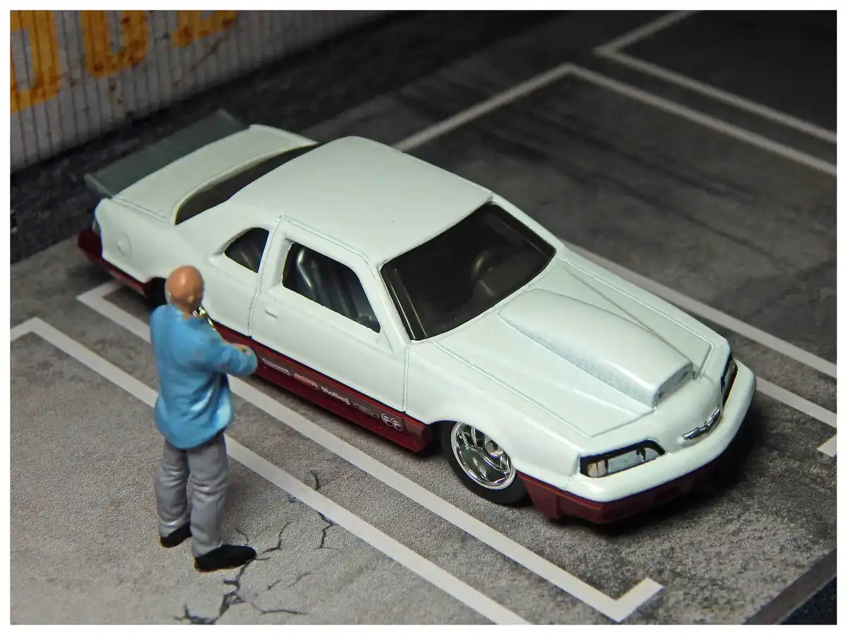A miniature figure of a bald man in a blue shirt and grey pants stands beside a white toy car with red trim and a large rear…