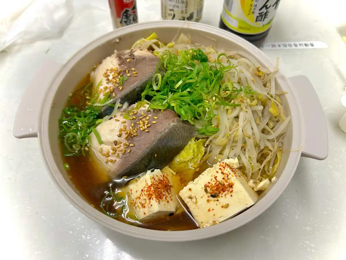 A close-up photo of a Japanese-style hot pot meal in a white ceramic bowl with handles, containing two large pieces of fish…