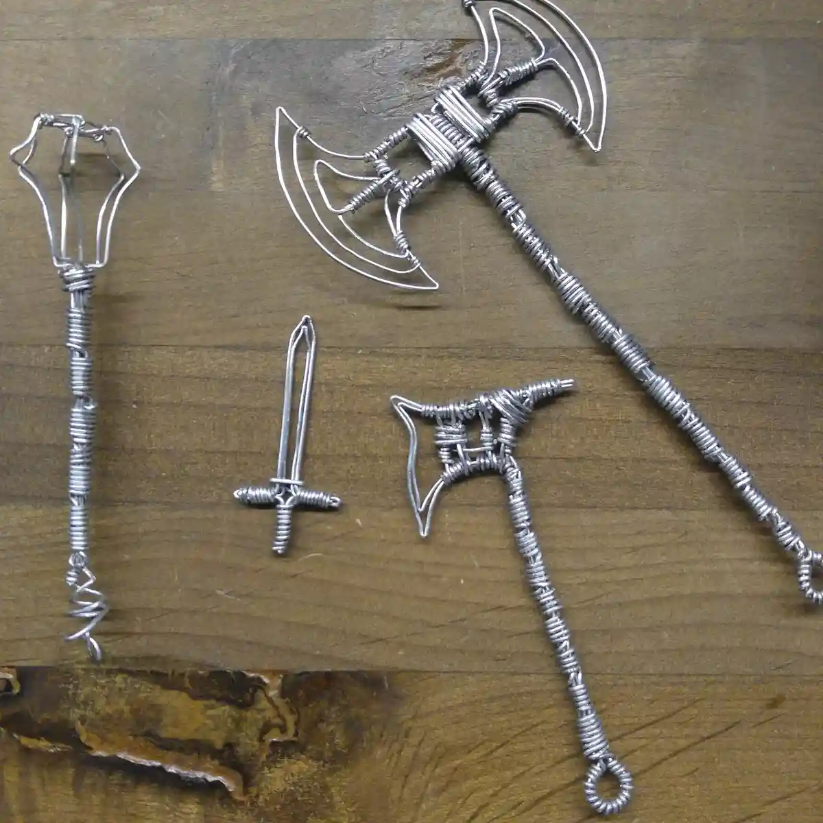 Four miniature wire sculptures of fantasy weapons — a double-headed axe, a sword, a throwing axe, and a mace — are arranged…