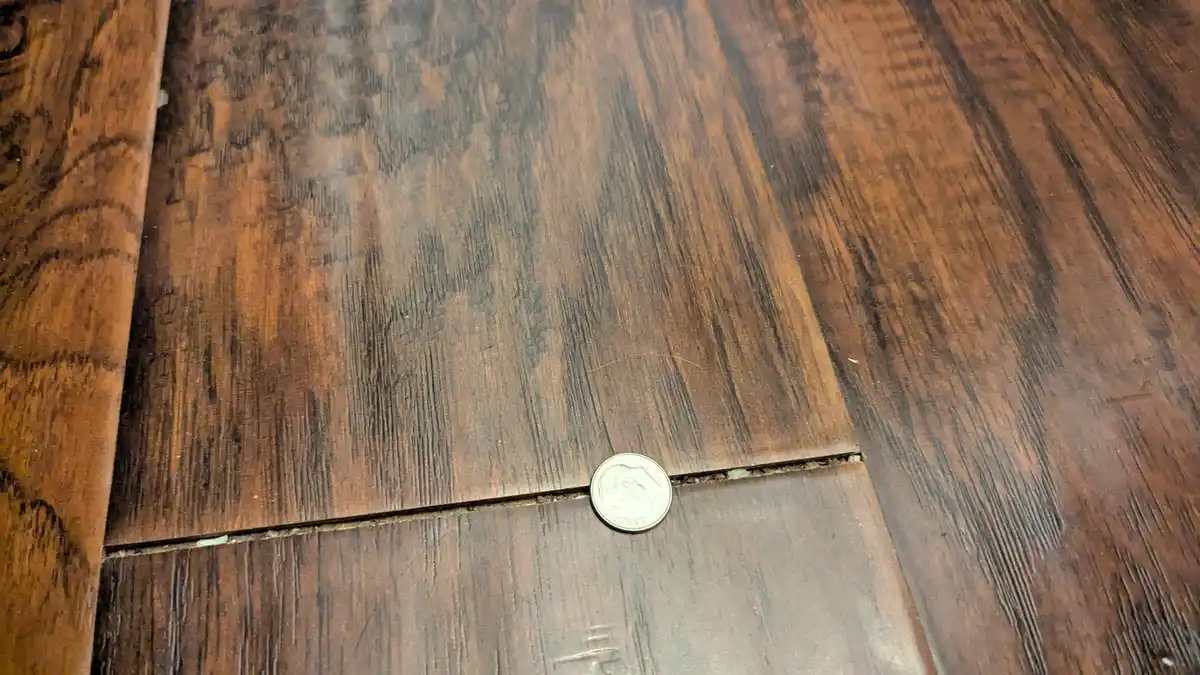 A close-up photo shows a U.S. quarter coin resting in the seam between two dark brown wooden floorboards, with visible gaps…