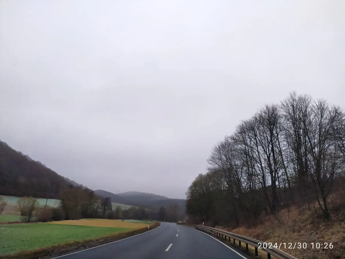 A photo of a rural road winding through a hilly, overcast landscape with bare trees on the right and green and yellow fields…