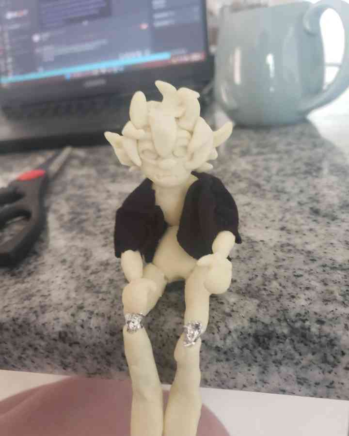 A handmade clay figurine of a character with spiky hair and a black jacket sits on a speckled countertop, held by a person's…