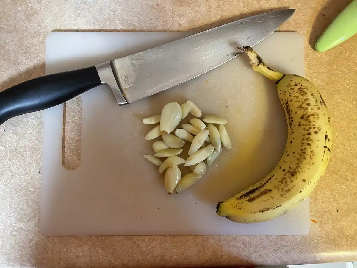 A top-down photo shows a ripe banana with brown spots and a pile of peeled garlic cloves on a white plastic cutting board. A…
