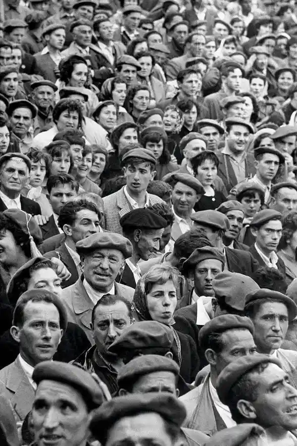 A black-and-white historical photo shows a dense crowd of people, mostly men wearing berets or caps, with varied…
