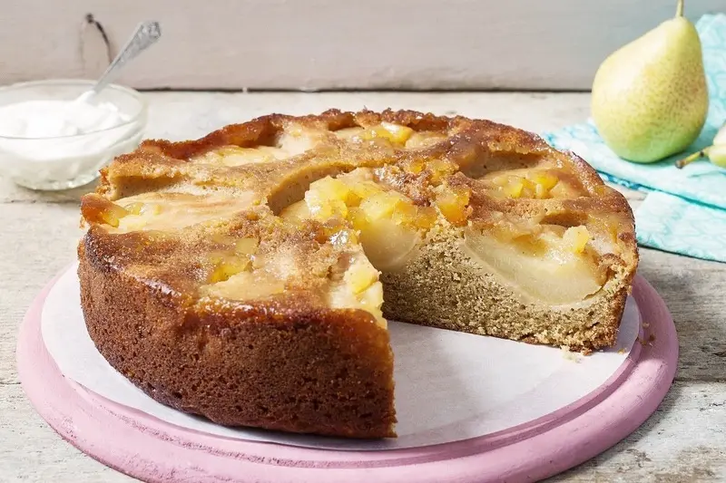 A golden-brown baked cake with visible chunks of fruit, likely pears, sits on a pink plate with a slice removed. In the…