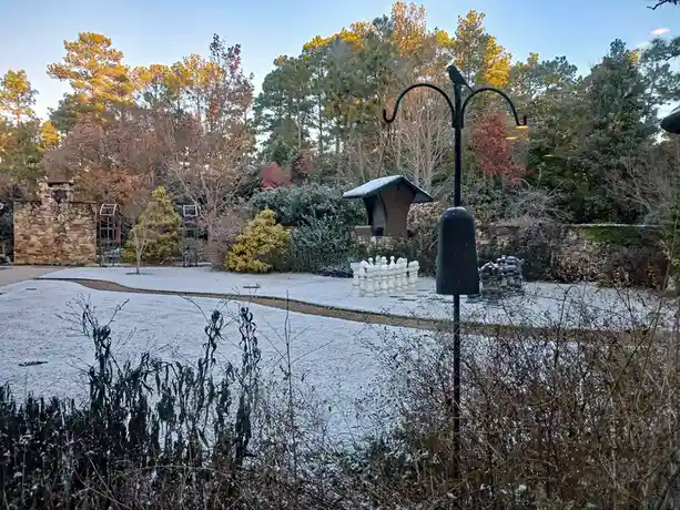 A snowy garden scene features large outdoor chess pieces arranged on a path, with a bird feeder and a decorative bird…