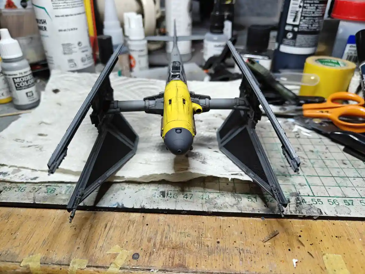 A partially painted Star Wars TIE fighter model sits on a cluttered workbench surrounded by modeling supplies like paint…