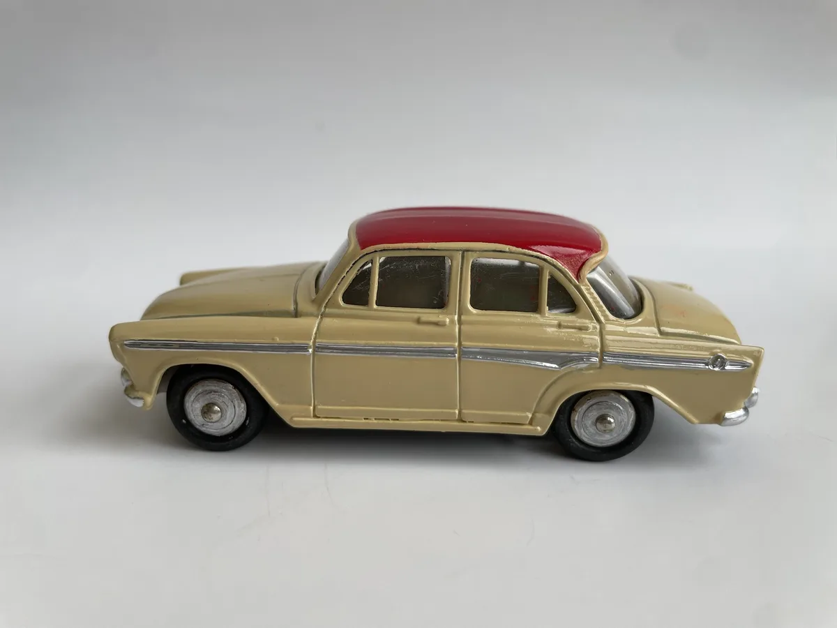 A vintage-style toy car with a beige body and a contrasting red roof is shown in profile against a plain white background…