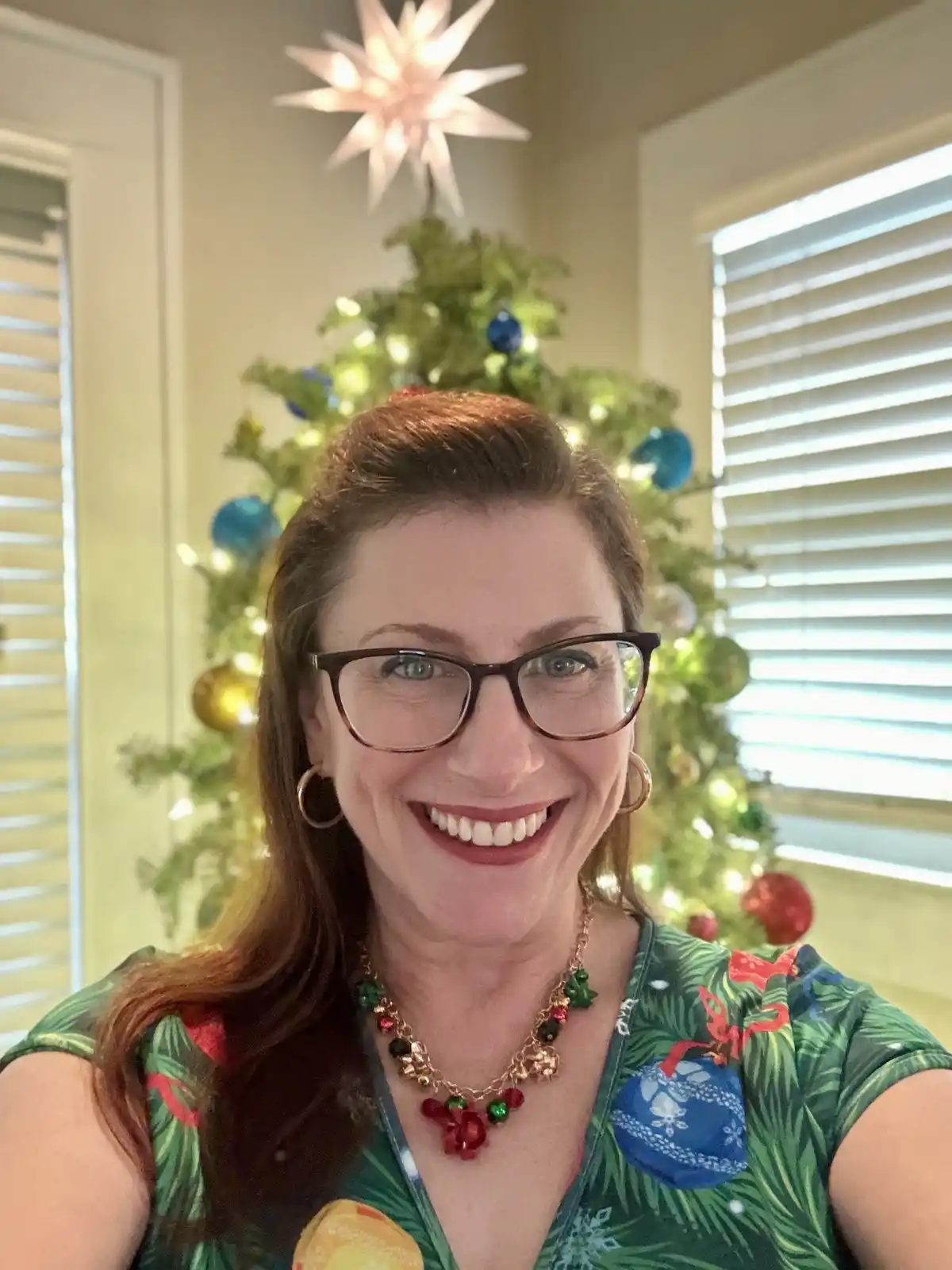 A woman with glasses and a festive green shirt featuring Christmas ornaments smiles at the camera, standing in front of a…