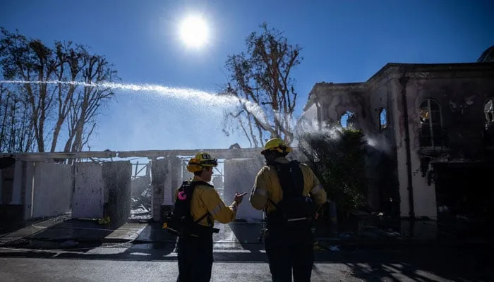 Two firefighters in yellow turnout gear stand in front of a severely burned house, spraying water from hoses under a bright…