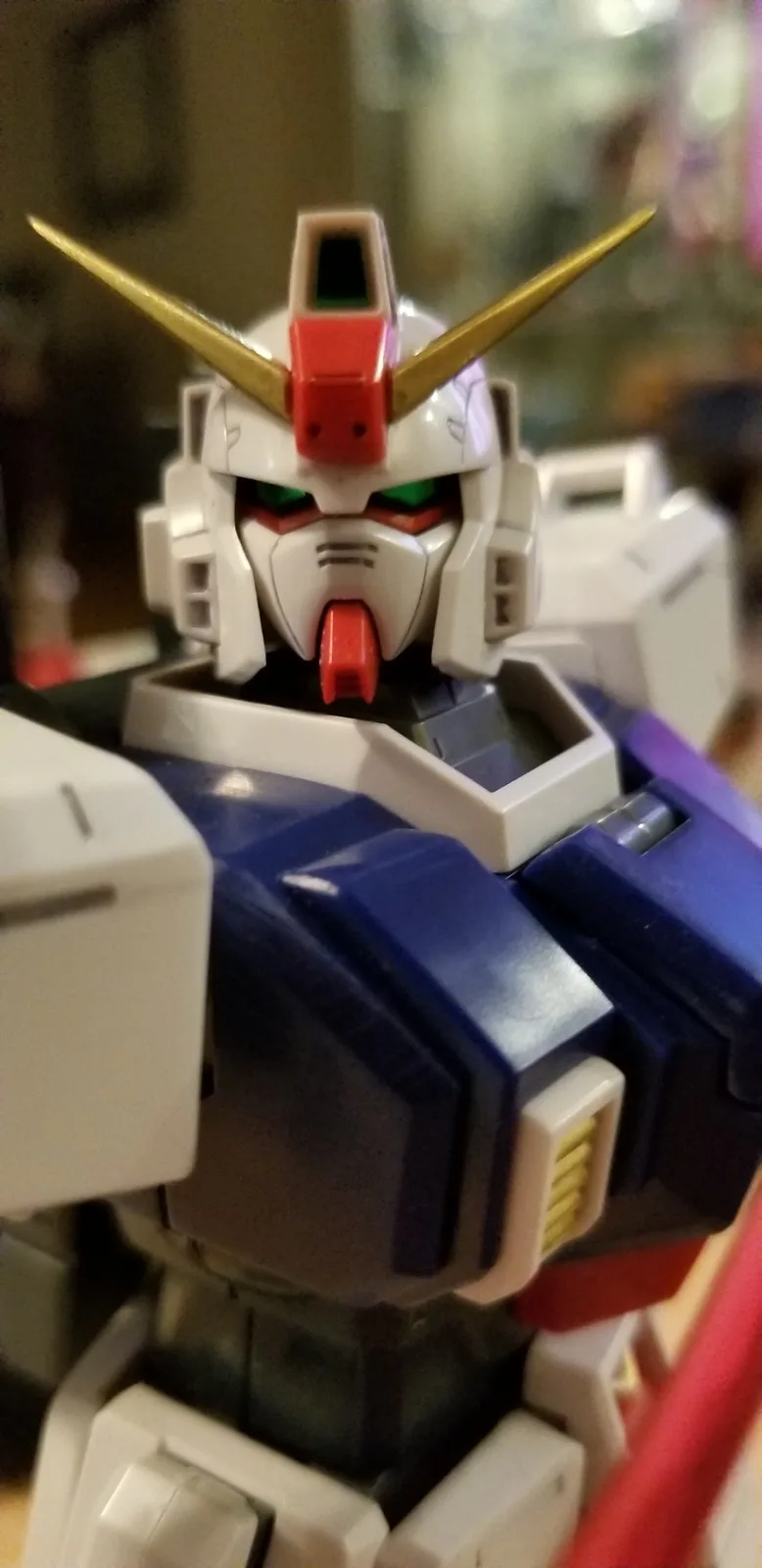A close-up photo of a white, blue, and red Gundam model toy with gold antennae and green glowing eyes, looking directly…