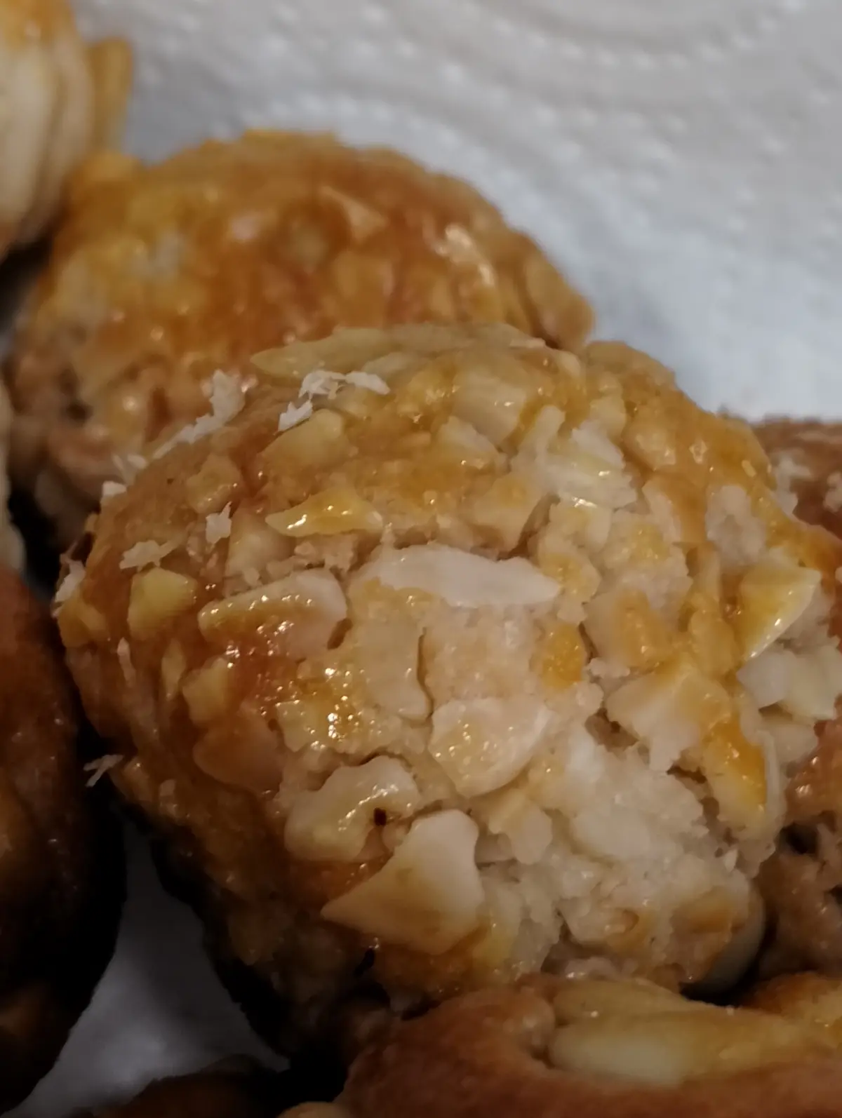A close-up photo of golden-brown, round cookies topped with chopped nuts and a glossy glaze, resting on a white paper towel…