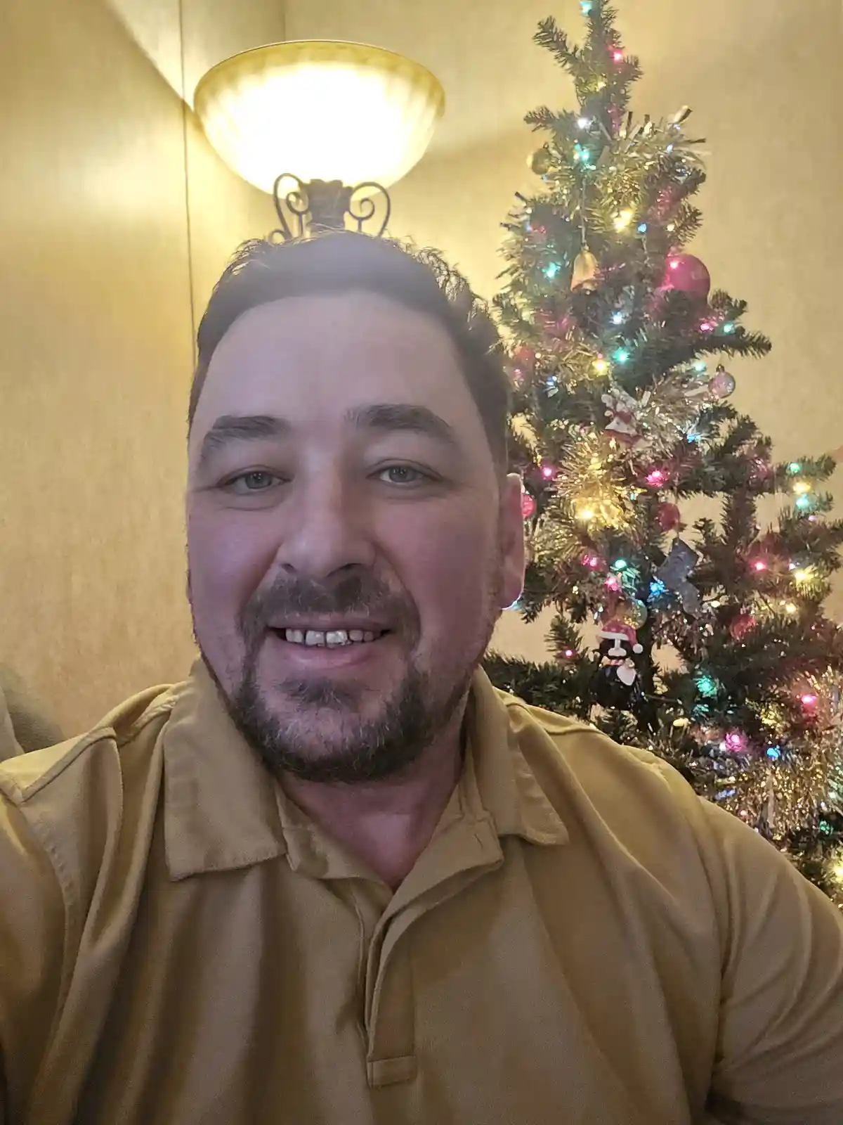 A man with a beard and mustache wearing a tan polo shirt smiles at the camera while standing in front of a decorated…
