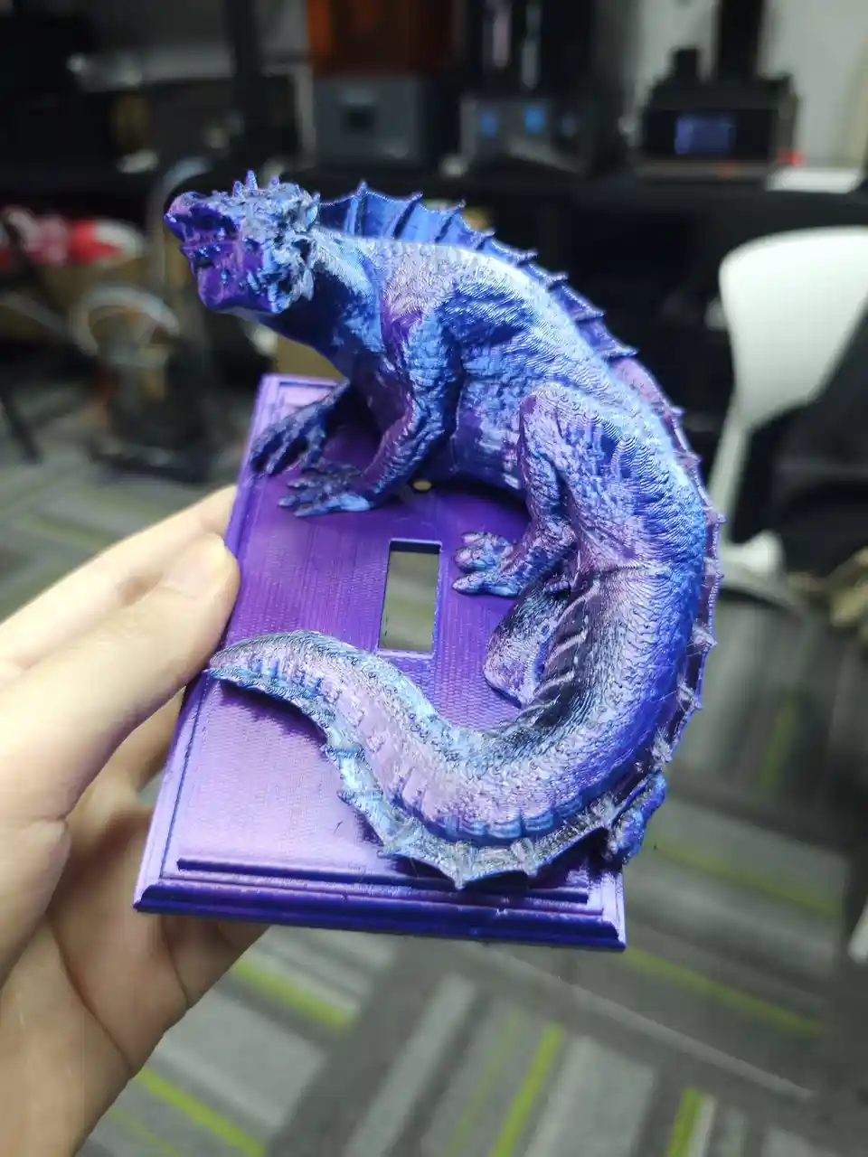 A hand holds a purple 3D-printed light switch cover shaped like a detailed, blue and purple dragon with spines and textured…