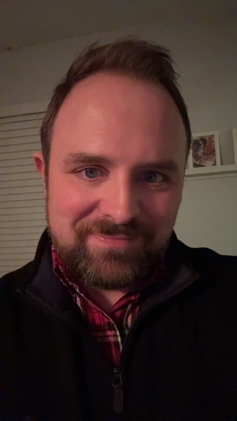 A man with blue eyes, a beard, and a receding hairline smiles at the camera while wearing a black zip-up jacket over a red…