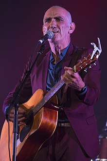 A bald man in a purple suit plays an acoustic guitar on stage under purple lighting, singing into a microphone. The image is…